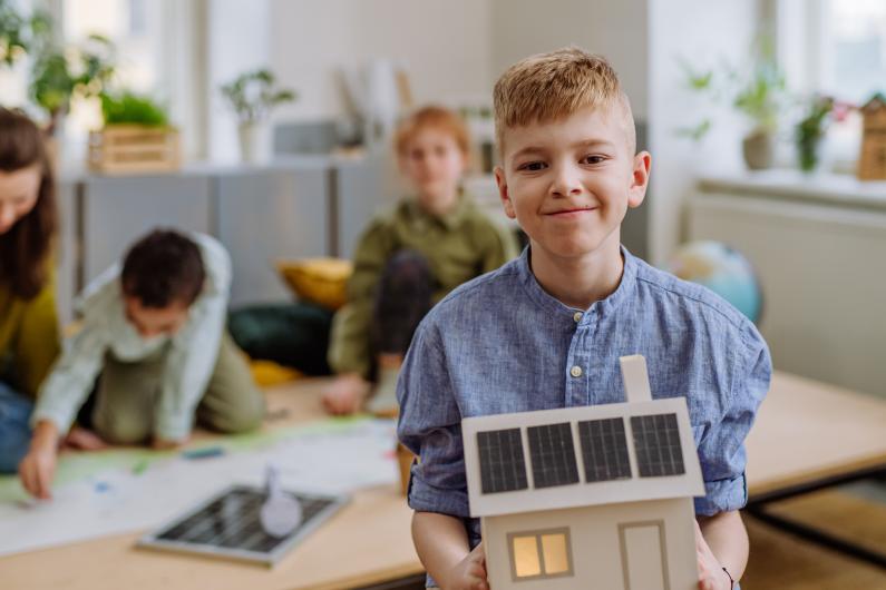 Petit garçon posant avec un modèle de maison avec système solaire pendant une leçon scolaire.